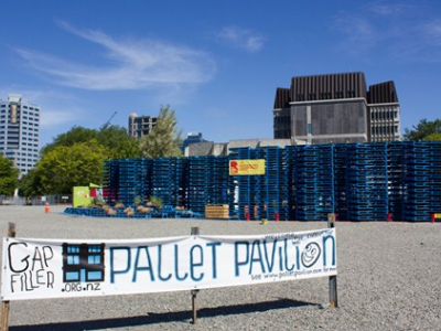 A structure made of pallets is in the background. In front a sign reads "Gap Filler Pallet Pavilion". In the distance two tall buildings can be seen, while in the foreground it is mostly gravel. 
